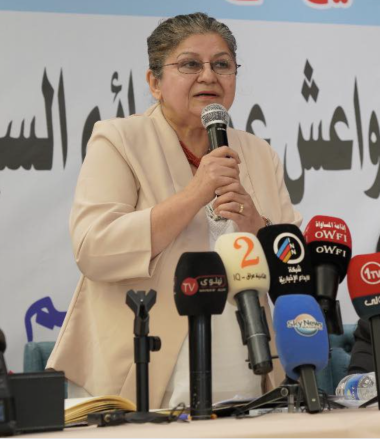 A woman wearing glasses and a light beige blazer (Yanar Mohammed) speaks into a handheld microphone at a press conference. She stands behind a table with several broadcast microphones in front of her, including ones labeled with TV channel logos. Arabic text is visible on the backdrop behind her.