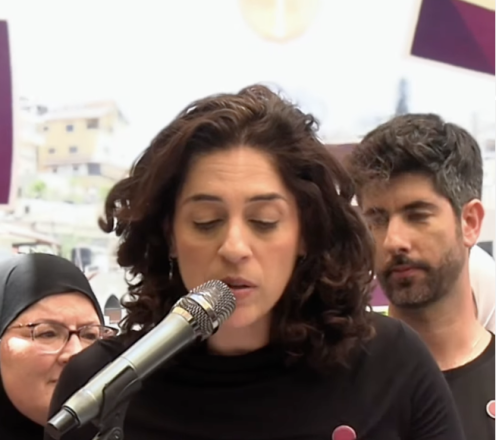 A woman with dark curly hair speaks into a microphone while reading from notes at a public event. Several people stand behind her, including a woman wearing a headscarf and glasses and a bearded man. The background contains blurred banners and display panels.