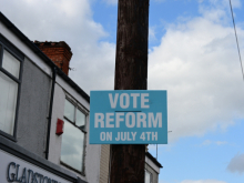 A blue campaign poster attached to a wooden pole reads 'VOTE REFORM ON JULY 4TH' in white bold letters. Part of a building with a sign reading 'GLADSTONE' is visible on the left, and the sky with clouds is in the background.