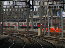 Railway junction with multiple electrified tracks, a London Underground train in the background, and a group of railway workers in orange high-visibility clothing walking beside the tracks.