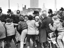 A group of women charging a police line. A colliery is visible on the horizon.