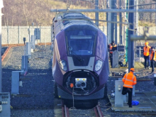 Purple East Midlands Railway train at a test or maintenance facility, with several railway workers in orange high-visibility clothing standing beside the tracks and equipment.