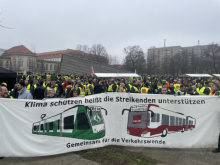 A large banner with a green tram, bearing the logo of the "Fridays for Future" climate strike group, next to a red bus bearing the logo of the Verdi trade union, with the slogan (in German): "Protecting the climate means supporting those on strike - Together for the transport transition."
