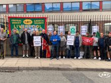 A group of RMT union members and supporters pose outside a railway facility, holding RMT “Official Picket” signs, red union flags and a large green-and-red “Central Mersey & North Branch Liverpool” banner. A red RMT flag, held reversed so it appears to read "TMR", is also visible. The group stands behind a pavement beside a fenced railway area.