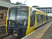 Yellow-and-black Merseyrail train standing at a station platform, displaying "Kirkby" as its destination, with a footbridge spanning the tracks in the background.