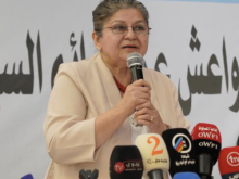 A woman wearing glasses and a light beige blazer (Yanar Mohammed) speaks into a handheld microphone at a press conference. She stands behind a table with several broadcast microphones in front of her, including ones labeled with TV channel logos. Arabic text is visible on the backdrop behind her.