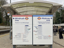 A London Overground station platform sign under a curved shelter. Two large panels show route maps: on the left, the Windrush line pointing left (Platform 2 via Whitechapel), and on the right, the Mildmay line pointing right (Platform 3 to Richmond and Clapham Junction). The signs include lists of stations with colored interchange symbols. Passengers stand on the platform to the right, and trees and railway infrastructure are visible in the background on an overcast day.