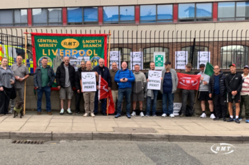 A group of RMT union members and supporters pose outside a railway facility, holding RMT “Official Picket” signs, red union flags and a large green-and-red “Central Mersey & North Branch Liverpool” banner. A red RMT flag, held reversed so it appears to read "TMR", is also visible. The group stands behind a pavement beside a fenced railway area.