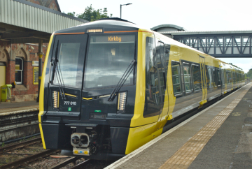 Yellow-and-black Merseyrail train standing at a station platform, displaying "Kirkby" as its destination, with a footbridge spanning the tracks in the background.