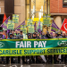 A group of trade union members stand in a city street holding a large green banner reading “FAIR PAY AT CARLISLE SUPPORT SERVICES” with RMT logos on either side. Several people behind them hold matching green and yellow placards with the same message, along with purple union flags and a red RMT flag. The group is gathered outside office buildings on a wet day, wearing coats and hats, appearing to take part in a pay protest or demonstration.