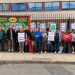 A group of RMT union members and supporters pose outside a railway facility, holding RMT “Official Picket” signs, red union flags and a large green-and-red “Central Mersey & North Branch Liverpool” banner. A red RMT flag, held reversed so it appears to read "TMR", is also visible. The group stands behind a pavement beside a fenced railway area.