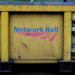 Close-up of a yellow industrial rail vehicle or equipment panel with the blue and red “Network Rail” logo painted on it, showing signs of wear, dirt, and metal fixtures along the edges.